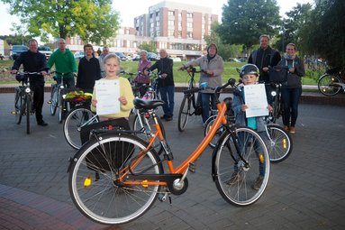 Ein Gruppe Menschen mit Fahrrädern, im Vordergrund ein oranges Fahrrad und zwei Kinder mit Urkunden