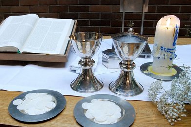Abendmahlstisch mit zwei Weinkelchen, zwei Teller mit Oblaten, eine helle Kerze und eine aufgelschlagene Bibel - Copyright: Andrea Enderlein
