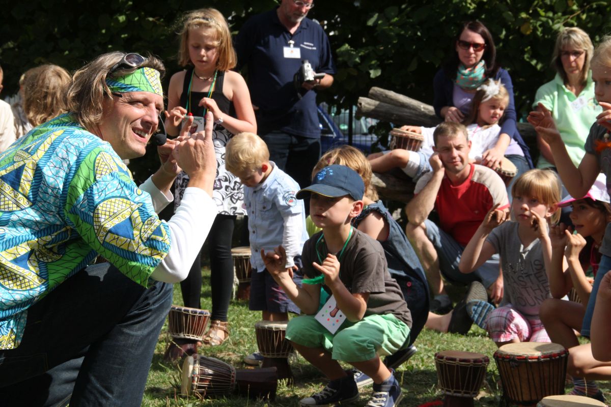 Viele Kinder hocken am Boden, ebenso ein grün gekleideter Mann und trommeln
