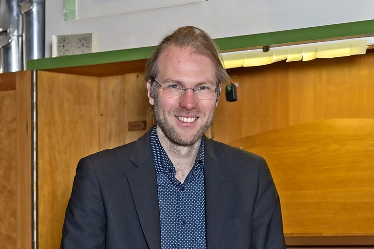 Junger Mann (Organist) sitzt mit dem Rücken zu einer Orgel und lächelt in die Kamera